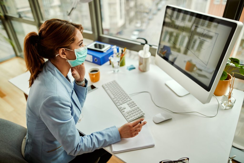 high angle view businesswoman with face mask analyzing blueprints desktop pc while working office
